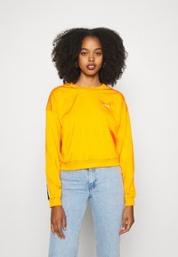 Felpa cropped arancione con scollo rotondo, maniche lunghe e dettaglio del logo. Abbinata a jeans azzurri chiari con un design minimale.