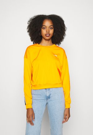 Felpa cropped arancione con scollo rotondo, maniche lunghe e dettaglio del logo. Abbinata a jeans azzurri chiari con un design minimale.