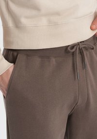 Brune sweatpants med elastisk talje og justerbar snøre. Side lommer og en glat, blød tekstur. Lysfarvet ribbet sweatshirt ovenfor.