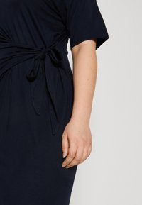 Navyblauwe jurk van zachte stof, met een knoopdetail bij de taille en korte mouwen, een gladde textuur en een vloeiende vorm.