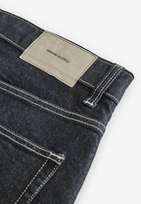 Sötétkék farmernadrág kontrasztos krém színű varrással, négyzet alakú bézs bőr címkével, amelyen a "DENIM SUPPLY" felirat szerepel, és egyszerű kialakítással.