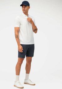 Weißes Poloshirt, marineblaue Kappe, dunkelblaue Shorts, weiße Sneakers mit beigen Akzenten und cremefarbene Socken. Tattoos auf dem linken Arm sichtbar.