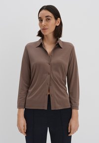 Chemise marron à manches longues avec col et coupe décontractée, présentant une texture lisse et un léger éclat, accompagnée d'un pantalon sombre.