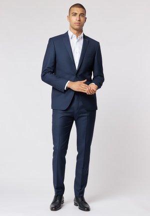 Abito blu navy con vestibilità slim, chiusura a un bottone, revers a punta e tasche frontali; abbinato a una camicia bianca e scarpe stringate nere.