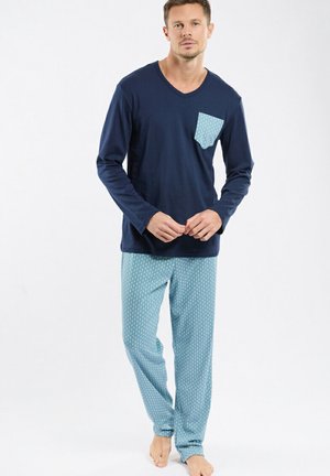 Homme portant un haut à manches longues bleu marine avec une poche poitrine à motif et un pantalon de pyjama assorti bleu clair à motif, debout pieds nus sur un fond blanc.