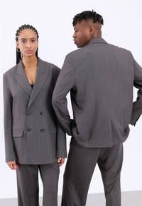Abito doppiopetto grigio a righe con ampi rever e tasche frontali, abbinato a pantaloni coordinati. Tessuto liscio con vestibilità strutturata.