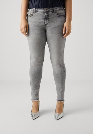 Person trägt ausgewaschene graue Skinny-Jeans mit ausgefransten Säumen und metallisch silberne spitz zulaufende High Heels, steht vor einem schlichten Hintergrund.