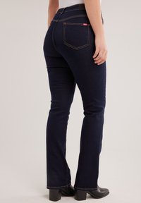 Žena u tamnoplavim trapericama ravnog kroja i crnim čizmama s potpeticom, stoji na svijetloj neutralnoj pozadini.