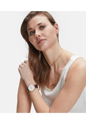 Braunem Lederarmbanduhr mit einem weißen Zifferblatt, ausgestattet mit silbernen Stundenmarkierungen und einem polierten Metallgehäuse. Das Model trägt eine silberne Kettenkette.