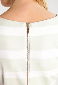 Pruhovaný svetr s mentolově zeleným a bílým vzorem, se zlatým zipem na zadní straně, hladkou texturou a širokým výstřihem.