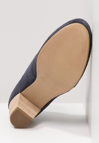 Granatowe zamszowe buty na drewnianym obcasie blokowym z teksturowaną podeszwą z falistym wzorem. But jest ustawiony pod kątem na jednolitym tle.