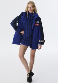Blaue Jacke mit mehreren Taschen, kontrastierenden schwarzen Ärmeln und Aufnähern. Getragen über einem blauen gemusterten Kleid, kombiniert mit schwarzen Sportschuhen.