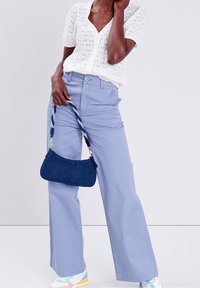 Sac en jean de forme courbe avec une bandoulière tressée, porté avec un haut texturé blanc et un pantalon large bleu clair. Des baskets colorées complètent le look.