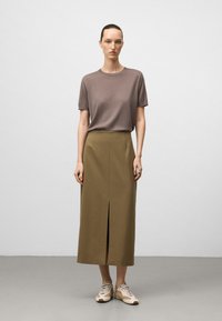 Donna in piedi con un top corto taupe a maniche corte, una gonna midi oliva con spacco frontale e scarpe da ginnastica beige e bianche, su uno sfondo grigio chiaro uniforme.