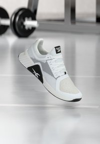 Λευκό και μαύρο αθλητικό παπούτσι Reebok που αιωρείται πάνω από το πάτωμα ενός γυμναστηρίου με θολούς αλτήρες και εξοπλισμό στο φόντο.