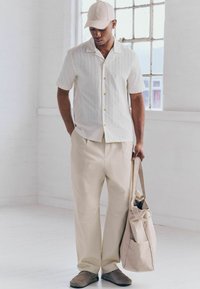 Lyst farvet kortærmet skjorte med knapper og lodrette striber, beige vidde-bukser, grå slip-on sko og en beige taske.