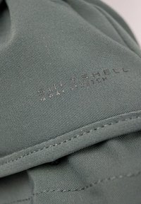 Weiches Softshell-Gewebe in mattem Grün, mit einer glatten Textur und einer sichtbaren genähten Naht. Der Markenname "SOFTSHELL 4 WAY STRETCH" ist geprägt.