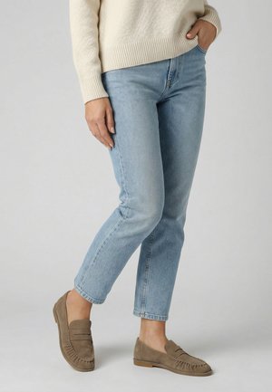 Žena stojící na šedém pozadí, má na sobě světlé modré džíny, béžový pletený svetr a taupe semišové mokasíny, s jednou rukou v kapse.