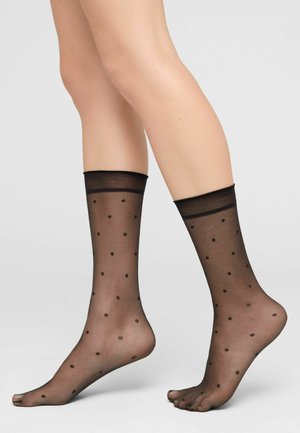 Jambes portant des chaussettes noires transparentes à la cheville avec de petits pois noirs, posées avec un pied à plat et l'autre légèrement relevé.