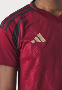 Červený sportovní dres s výstřihem do V, černými akcenty na ramenou a texturovaným diamantovým vzorem. Zlaté logo Adidas na hrudi.