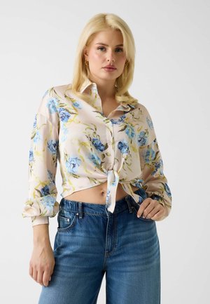 Młoda kobieta o blond włosach nosi związaną kwiecistą bluzkę z niebieskimi kwiatami i wysokimi niebieskimi dżinsami, pozując na jednolitym tle.