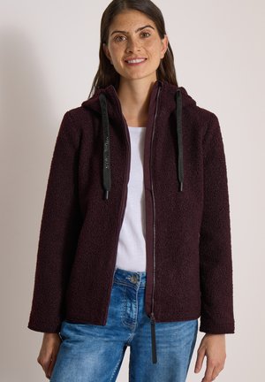 Mujer con sudadera con capucha con cremallera texturizada de color púrpura oscuro y cordones negros, camisa blanca y vaqueros azules, de pie frente a un fondo liso.