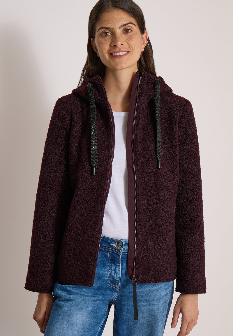 Femme portant un sweat à capuche zippé texturé violet foncé avec des cordons noirs, une chemise blanche et un jean bleu, debout devant un fond uni.