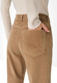 Pantalons en velours côtelé marron avec une finition texturée, dotés d'une poche arrière et d'une taille élastique. Haut crème à côtes partiellement visible.