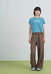 Kortärmad blå t-shirt med text och breda bruna rutiga byxor med elastisk midja; modell i flerfärgade sneakers; minimalistisk bakgrund.