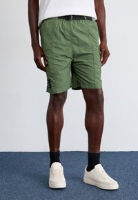 Žalios spalvos lengvi šortai su elastine juosmens juosta, šoninėmis kišenėmis ir juodu etiketės detaliu. Derinami su baltais sportbačiais ir juodomis kojinėmis.