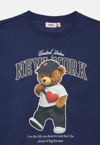 Tumit blu džemperis ar uzdrukātu teddy lāci baltā kreklā ar sarkanu sirdi un melnām biksēm, teksts virs un zem grafikas.