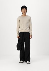 Beige strikket sweater, sorte brede bukser, grå og hvide sko og en lille sort skuldertaske. Ensfarvet baggrund.