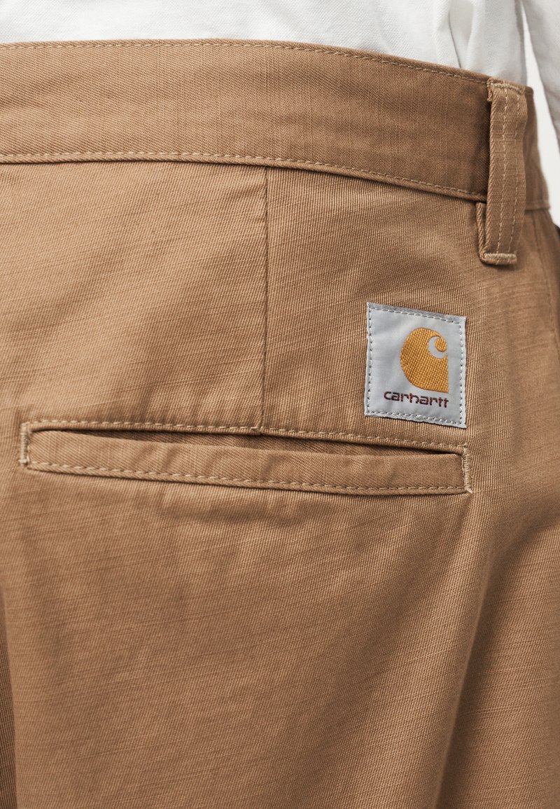 Tan woven trousers with a back pocket featuring a branded label. The fabric has a smooth texture and a clean finish.