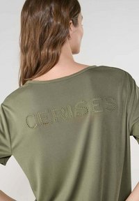 Camiseta verde oliva confeccionada en material suave, con la inscripción bordada en letras recortadas "CERISES" en la parte posterior en un diseño redondeado y casual.