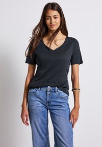 T-shirt noir à col en V avec des manches courtes, fabriqué dans un tissu lisse. Associé à un jean large bleu clair et à un bracelet en or au poignet.