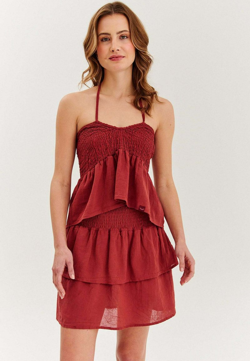 Žena v bezrukávových, tierovaných červených šatách so zmršteným živôtikom a halter krkom, stojí proti jednoduchému svetlému pozadiu.