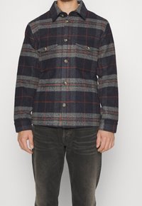 Man wearing a dark plaid button-up jacket with chest pockets, paired with black faded jeans, standing against a plain light background.