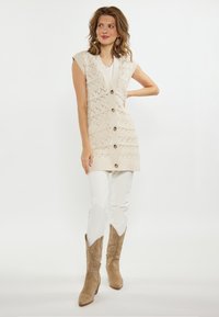 Beige gebreide vest met een V-hals, korte mouwen en ruitpatronen aan de voorkant, met houten knopen. Witte broek en tan cowboylaarzen.