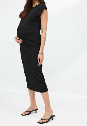 Zwangere vrouw in een zwarte midi-jurk met structuur en zwarte sandalen met hak, zijwaarts staand met één hand op haar buik.