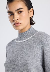 Jeune personne aux cheveux courts et foncés, portant un pull en tricot gris avec une rayure blanche près du col et une petite boucle d'oreille en or.