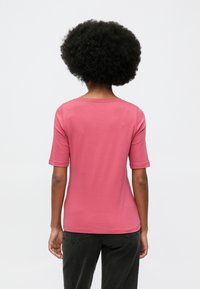 Άτομο με αφρο χτένισμα, φορώντας ένα απλό ροζ κοντομάνικο πουκάμισο και μαύρο παντελόνι, στέκεται με την πλάτη σε λευκό φόντο.