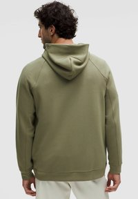 Un homme aux cheveux noirs bouclés portant un hoodie vert olive et un pantalon blanc, tourné le dos contre un fond clair.