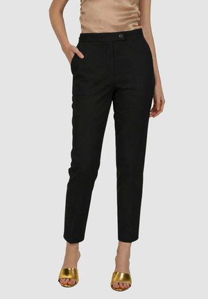 Donna che indossa pantaloni neri aderenti fino alla caviglia e tacchi aperti dorati, in piedi con una mano in tasca.