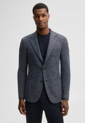 Man die een donker textuurde blazer draagt over een zwart overhemd, staand met handen ontspannen langs zijn zij tegen een effen lichte achtergrond.