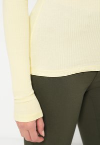 Vaaleankeltainen ribbikuvioinen pitkähihaisen paita, jossa on istuva muotoilu ja peukaloreijat, yhdistettynä tummanvihreisiin joustaviin housuihin. Pehmeä rakenne ja yksinkertainen ommel.