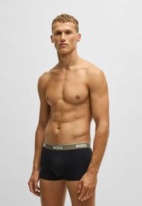 Schwarze Boxershorts aus einem weichen, dehnbaren Material mit einem breiten olivgrünen Bund, auf dem der Markenname "BOSS" in Weiß aufgedruckt ist. Figurbetontes Design.