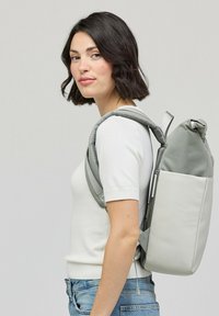Zaino grigio realizzato in materiale morbido, con una parte superiore arrotondata, tasca laterale e spallacci regolabili. Indossato sulla schiena, abbinato a una camicia leggera.