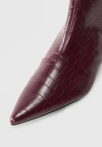 Stivale alla caviglia bordeaux con punta affusolata, realizzato in materiale sintetico lucido con un motivo a coccodrillo in rilievo e un design elegante.