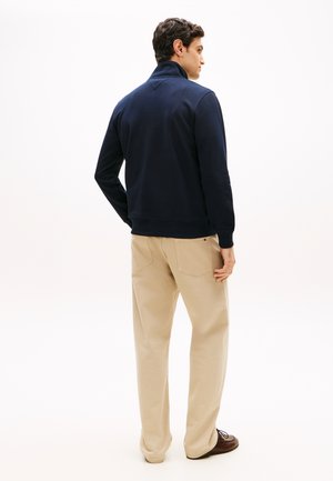 Homme debout de dos, portant une veste bleu marine, un pantalon beige et des chaussures marron sur un fond clair uni.