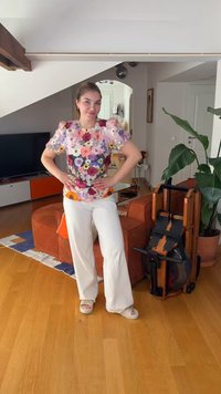 Femme debout, souriante, les mains sur les hanches, portant un haut fleuri et un pantalon blanc dans un salon avec un rameur en bois et une grande plante verte.